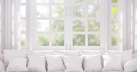 White couch with multiple throw pillows in front of large windows with sheer curtains. Bright, airy interior setting with natural light filtering through.