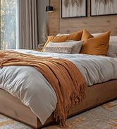 Bedroom interior featuring bed with orange throw blanket and tan pillows against white wall.